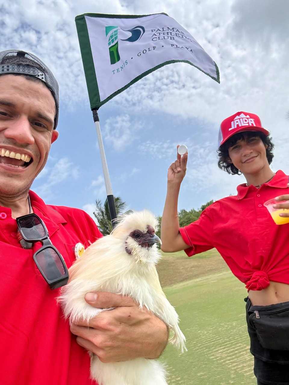 Armando, cofundador de punto.pio, sonriendo mientras sostiene una gallina Silkie junto a la bandera de golf de Palmas Athletic Club, durante SME Golf